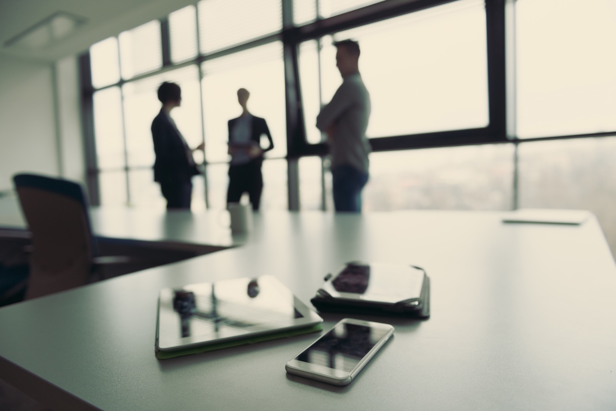 Close up of smart phone and tablet computer at office meeting room. Business people group interacting in background.